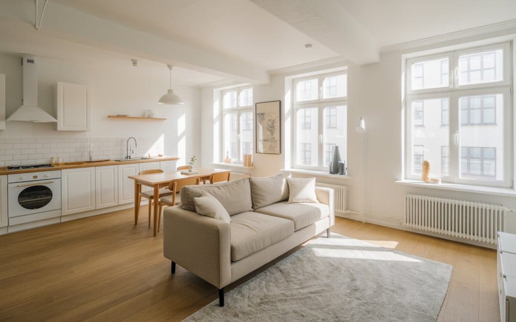 Apartment living room and kitchen with white walls, beige sofa, light gray rug, natural daylight streaming through large windows, cozy minimal aesthetic, Pinterest-worthy interior
